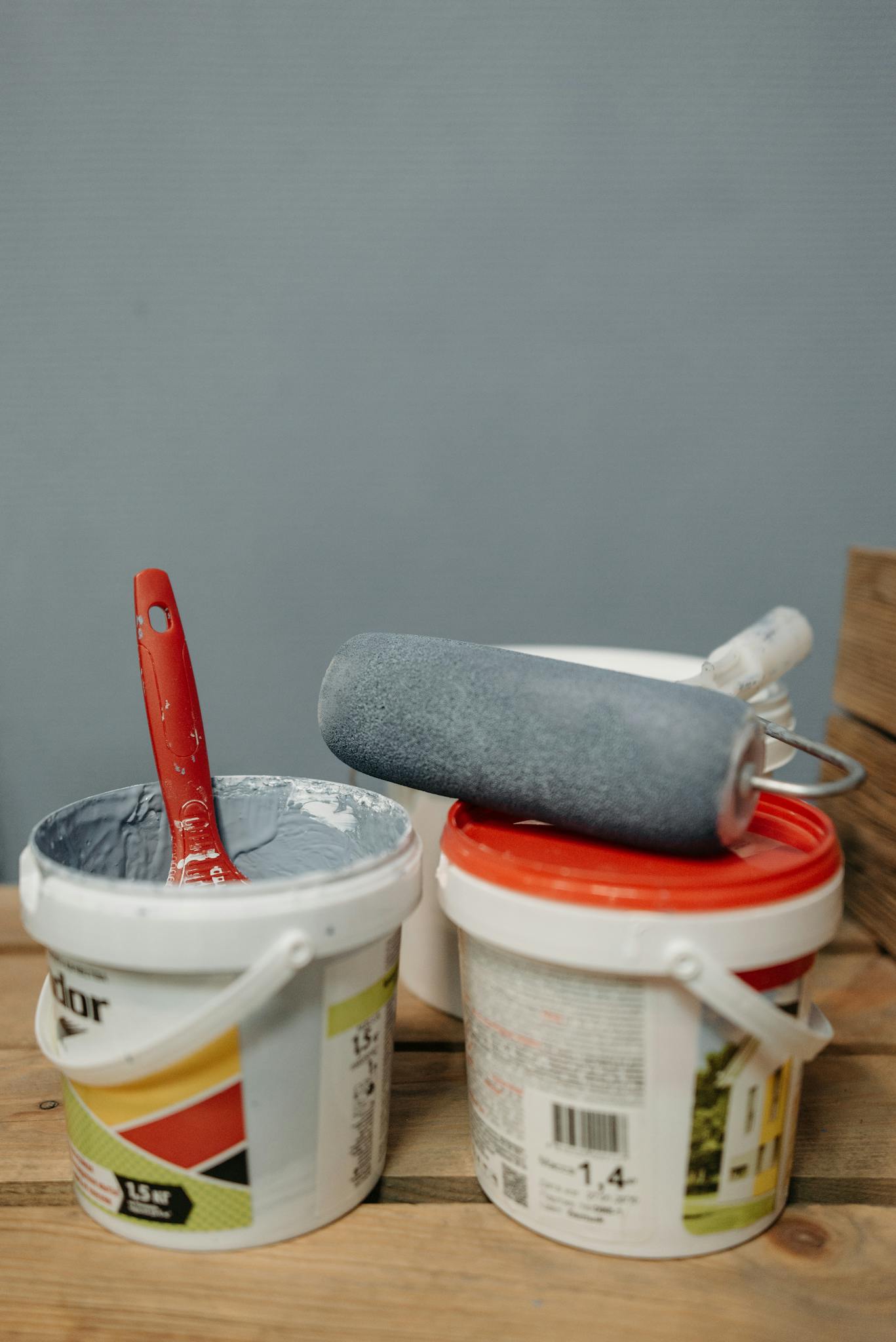 Close-up of paint buckets with brushes and roller on wooden surface for decorating projects.
