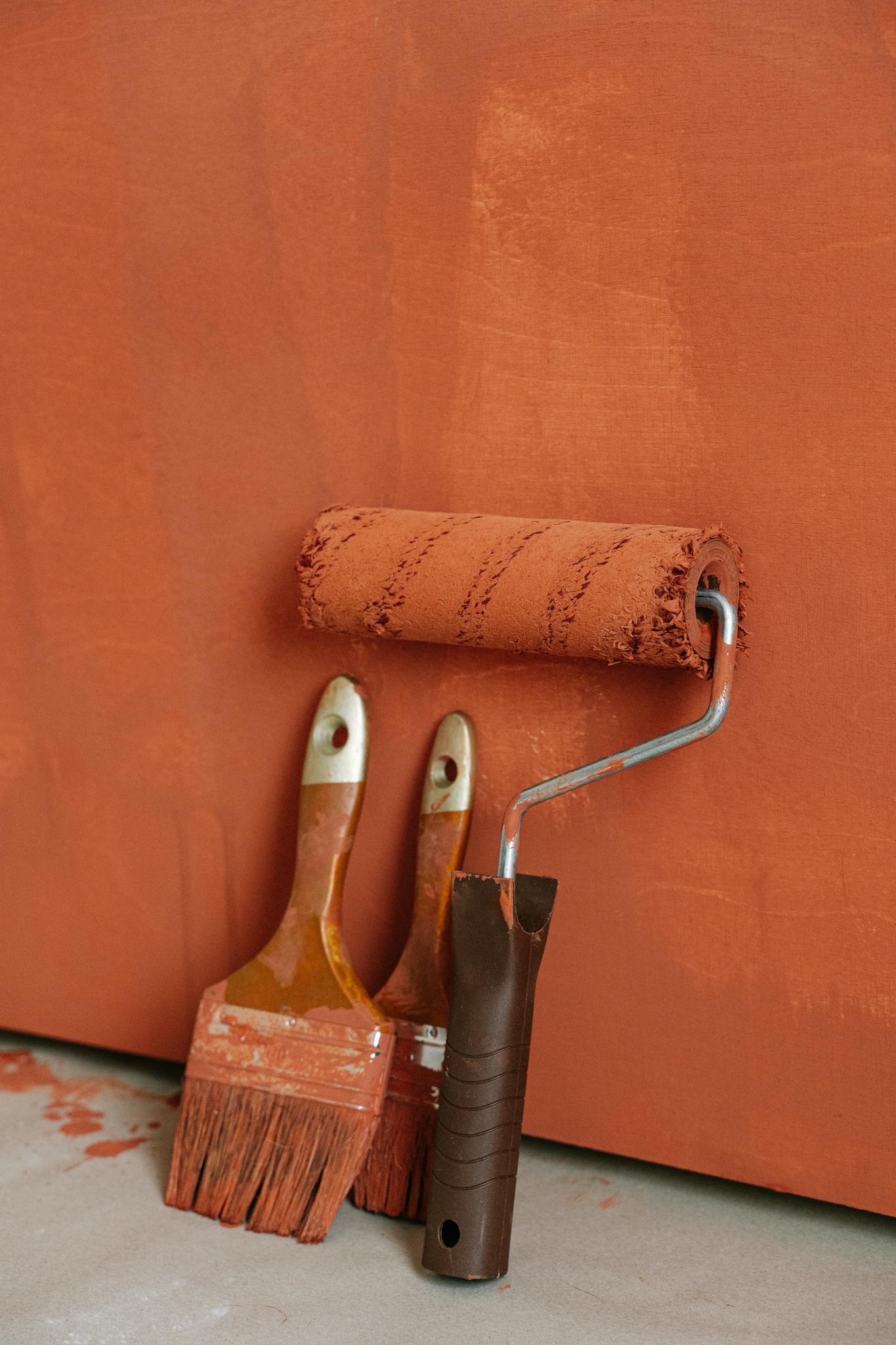 Three paintbrushes and a roller with red paint are placed against a freshly painted wall, showcasing DIY painting tools.